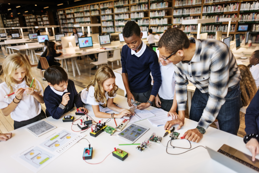Students working on electronics projects with a teacher in a STEM lab filled with computers and bookshelves.