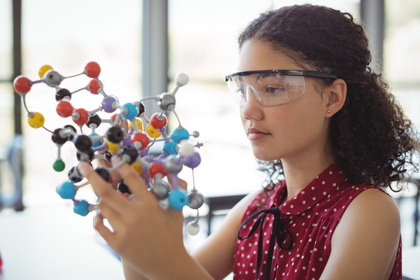 Teen girl in safety goggles examining a colorful molecular model in a STEM lab setting.