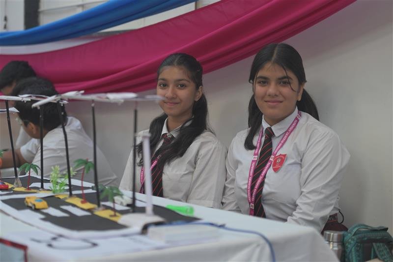 Two schoolgirls with a smart city model at a STEM exhibition booth.