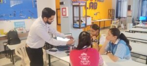 A teacher mentoring schoolgirls in a STEM Lab in Vadodara, explaining an electronic component at a workstation with tools in the background.