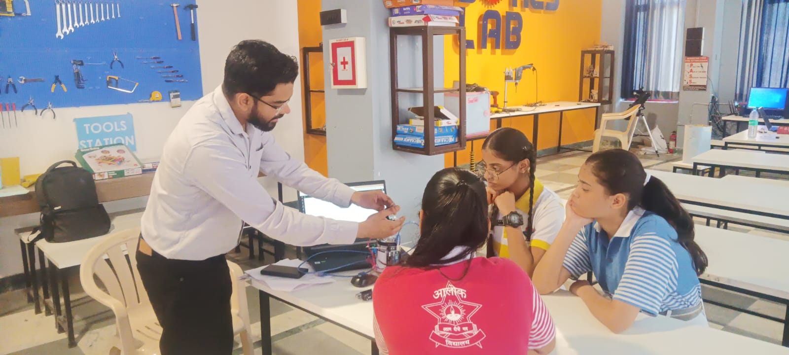A teacher mentoring schoolgirls in a STEM Lab in Vadodara, explaining an electronic component at a workstation with tools in the background.