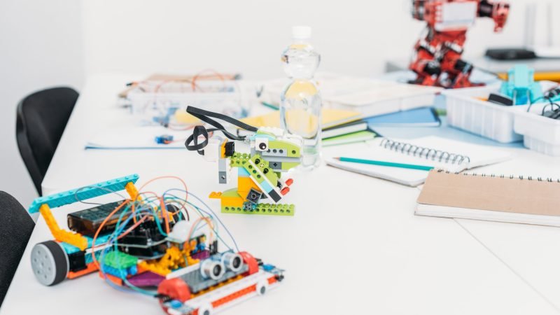 Various colorful DIY robot models made from LEGO and electronic components displayed on a white table in a classroom or workshop setting.